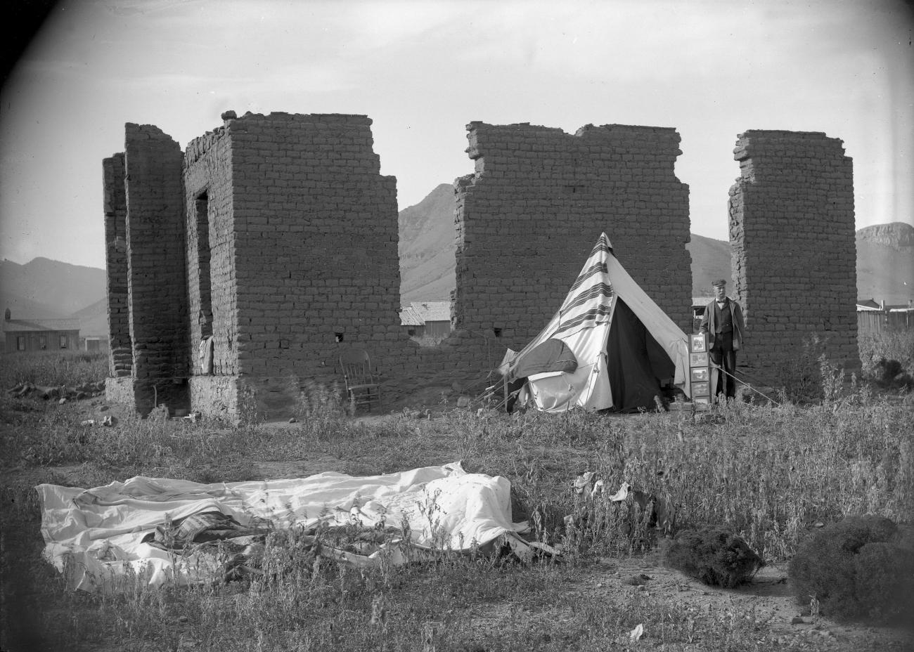 Walter Johnson stands next to ruins of the Grand Hotel in Magdalena, New Mexico. A rack of photographs is visible beside him. September, 1898.