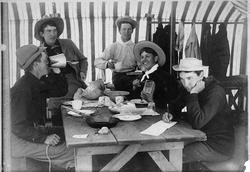 Five men in hats sit at a table having breakfast. 