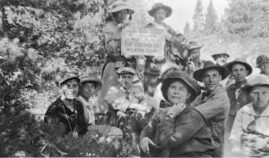 A group of men and women pose in the Clear Creek Basin region. Most of them are sitting on the ground, but two women stand in the back with a club sign.