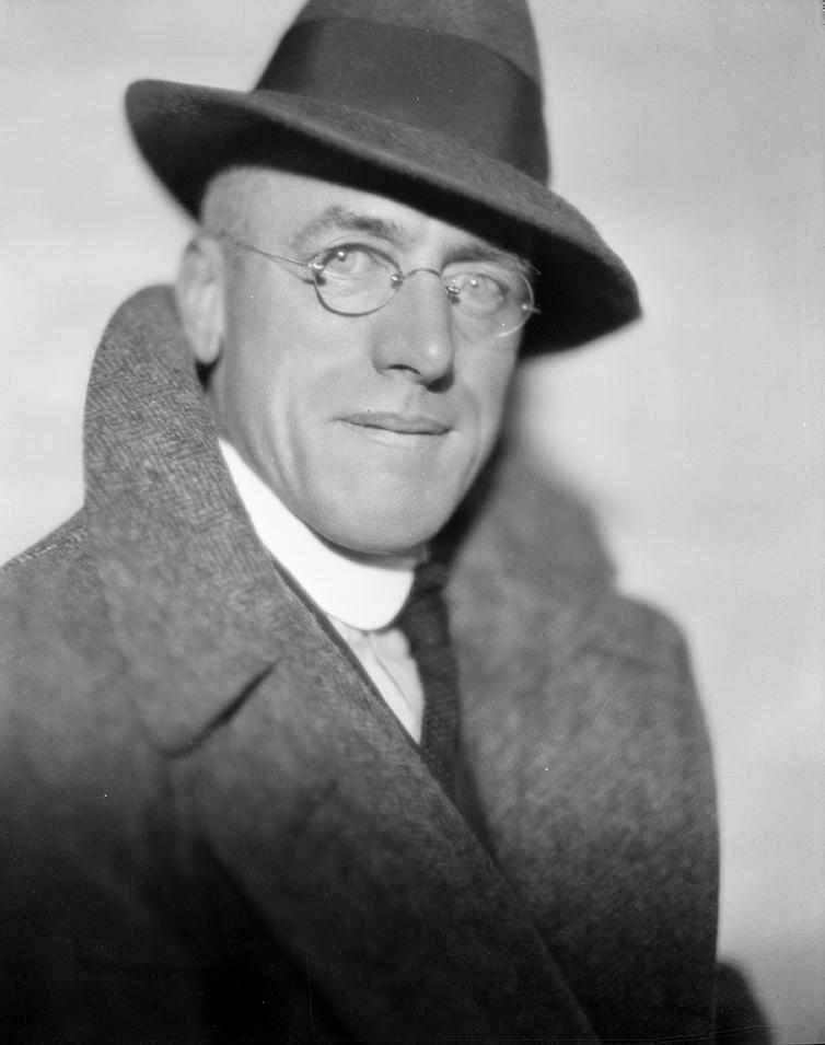 Fred Payne Clatworthy looking debonair, staring into the camera, wearing glasses, a hat, and a peacoat with popped collar.