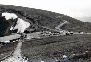 A black and white photo of the road with a parking lot in the center of the picture next to a structure and on the other end rolling into the hills in the distance. 