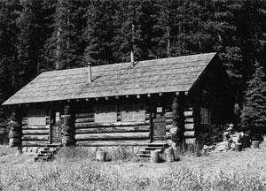 A black and white photo of the cabin with two doors and stairs leading up to them beneath overhanging roof. In the background are a wall of evergreen trees. 