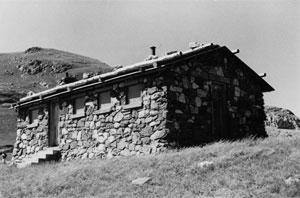 A black and white photo of the cabin with stone walls, the door with a series of steps lead up to it. 