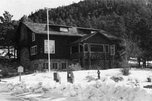 A photo of the lodge in black and white with snow in the foreground and forested mountains in the back. 
