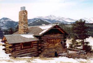 A view of the cabin with log walls and gabled roof and large stone chimney on the side in front of mountains in the distance. 