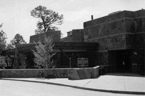 A black and white photo of the building with covered path and trees sticking up sporadically on the left side. 