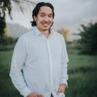 Photo of author Garrett Briggs, a younger man standing in a green field with trees in the background.  He has collar length black hair which is parted in the middle of his head and frames his face. He is relaxed, with his fingertips tucked into the front pockets of his khaki pants, and his light colored dress shirt unbuttoned at the neck.  He has a faint black mustache and goatee, and is smiling.