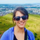 Photo of Kristin Chiesi. She has longer dark hair that is pulled up, and is wearing sunglasses, a black top, and a blue hoodie jacket. She is standing on a hilltop, and behind her is a