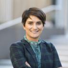 Photo of professor Samira Rajabi. She is standing outside of a building, the steps of which can be seen in the distant background. Samira is standing with her arms crossed, smiling. She has short dark hair, and is wearing a dark green and dark blue plaid blazer with a green and blue lacy blouse that has a high lacy collar.