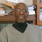 Photo of professor William B. Allen. He is an African American man, seated in a room with many shelves behind him full of books of all sizes. He is smiling. He is a bald man, with some salt and pepper short facial hair and he wears eyeglasses.  In this photo he is wearing a gray v-neck sweater with a black turtle neck shirt underneath it.