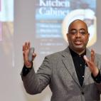 Photo of an African American man standing in front of an unseen audience. He is speaking, and his hands are in the air as he is describing something. In his right hand, he appears to be holding a clicker, presumably to advance the images that are shown on the screen behind him. In this photo, those images are of the two books this author has written.