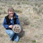 Photo of Bonnie Clark. She is smiling, and kneeling in a grassy field, holding a a round, gray object in her hands. The object has 8 slits in the bottom, as if to strain the contents of the vessel. 