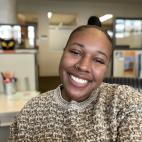Photo of Jordan Lewis, a young Black woman smiling for a close-up photo. She has hair that is pulled back tightly, and she does not wear eyeglasses or have any jewelry showing. She is wearing a neutral-colored, horizontal striped crew neck sweater She is sitting in a room with lightly colored walls and a window in the distant background. She is smiling widely for the camera.
