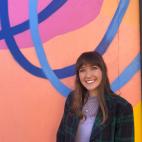 Photo of a young woman with long dark hair and bangs. She is smiling broadly while standing in front of a portion of very colorfully painted outdoor mural. The wall is orange and yellow, with a pink post and blue circles. She is wearing a purple turtle neck shirt, with a dark blue and green plaid coat that is unbuttoned in front, and a silver and pearl pendant around her neck.