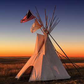 A tipi at sunset. Over the tipi fly two flags: the flag of the United States, and the white flag of peace.