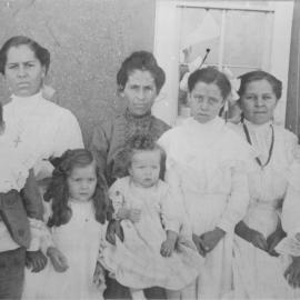 Four women and four children sit in front of an adobe house facing the camera in a black and white photograph
