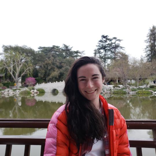 Photo of a young woman named Ariel Schnee, in a red puffy jacket, standing on a bridge with a lake and beautiful gardens in the background. She has long dark hair which sweeps to her right shoulder, rosy cheeks, and a wide smile.