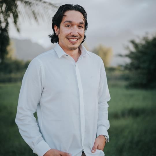 Photo of author Garrett Briggs, a younger man standing in a green field with trees in the background.  He has collar length black hair which is parted in the middle of his head and frames his face. He is relaxed, with his fingertips tucked into the front pockets of his khaki pants, and his light colored dress shirt unbuttoned at the neck.  He has a faint black mustache and goatee, and is smiling.