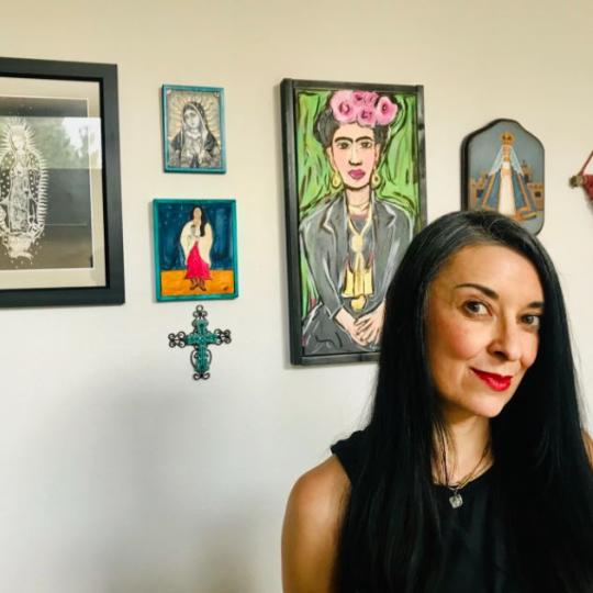 Photo of a woman standing in front of a white wall on which hangs several brightly colored images of the Virgin Mary, artist Frida Khalo, an ornate turquoise cross, another image of the Virgin Mary in a bright turquoise frame, and a macrame wall hanging made of crimson red yarns. The woman is dressed in a black dress and has long black hair with subtle stipes of gray on the top of her head.
