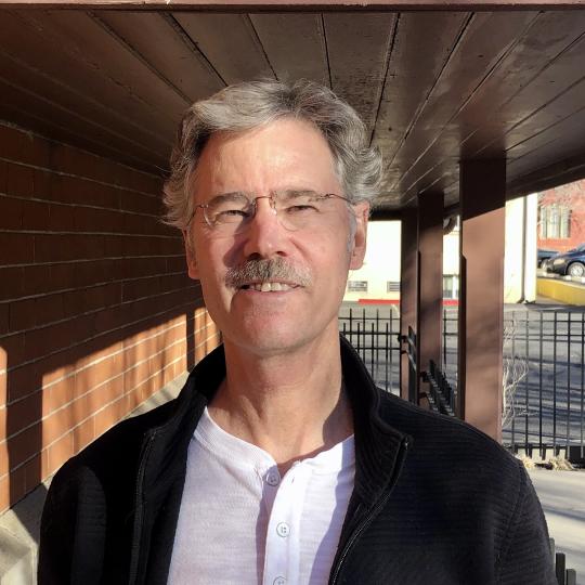 Photo of Kim Grant. He is wearing a black casual jacket and a white 3-button shirt. He has salt and pepper colored wavy short hair and wears glasses. He also has a moustache. He is standing on the porch of a building, as the roof overhead can be seen as well as the pillars supporting the porch roof.
