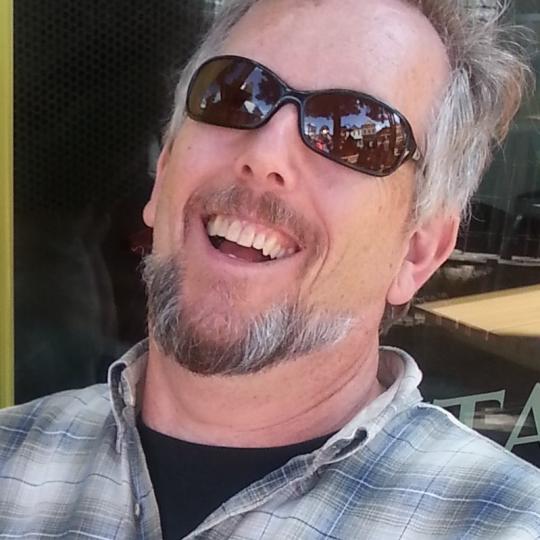 Photo of author Phil Carson. He is wearing a blue and gray plaid button-down collar shirt over a black crew neck t-shirt.  He has light brown-to-gray colored hair and a beard and mustache of the same.  He is pictured here on a bright day, wearing sunglasses and seems to be laughing.