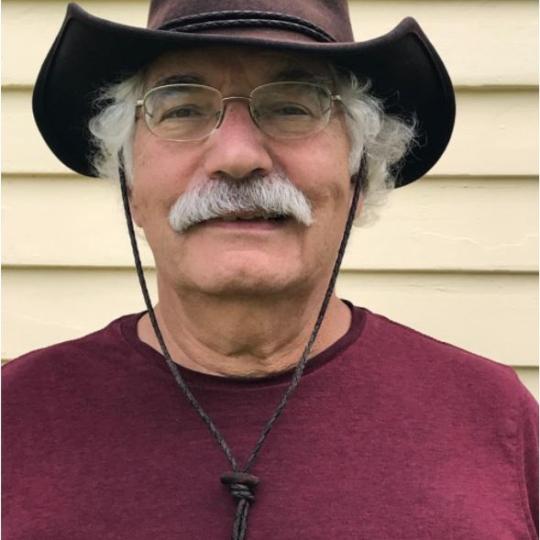 Photo of Robert Moll. He is standing in front of a building with yellow painted siding. Robert is wearing a burgundy colored t-shirt, with an outback-style dark leather hat. The had has a curled up brim on each side of his head, and there are cords from each side of the hat so that it can be tightened up under his chin. He has white curly short hair and a white moustache, and wears gold metal framed eyeglasses.