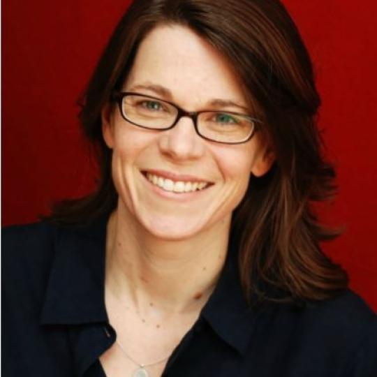 Photo of professor Susan Schulten. This image is a head shot, showing Susan in front of a red plain background. She is has long reddish-brown hair that is parted on her right side, and she wears rectangle-shaped eyeglasses. She is pictured here wearing a black button-up shirt and a silver pendant.