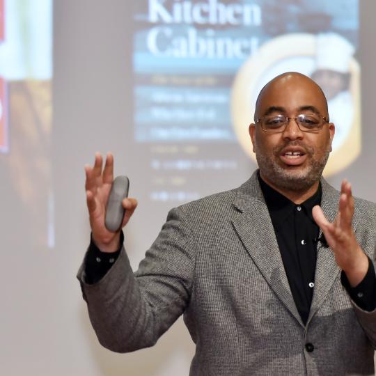 Photo of an African American man standing in front of an unseen audience. He is speaking, and his hands are in the air as he is describing something. In his right hand, he appears to be holding a clicker, presumably to advance the images that are shown on the screen behind him. In this photo, those images are of the two books this author has written.