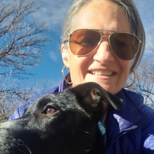 Photo of Cindy Nasky, with a dog. She looks to be hugging the dog, while the dog is looking off to its right and Cindy is looking at the camera. She is wearing a lightweight blue Patagonia jacket and aviator-style sunglasses Her hair is pulled back away from her face, and she is smiling. It is a sunny day, and the blue sky and treetops are visible behind her.