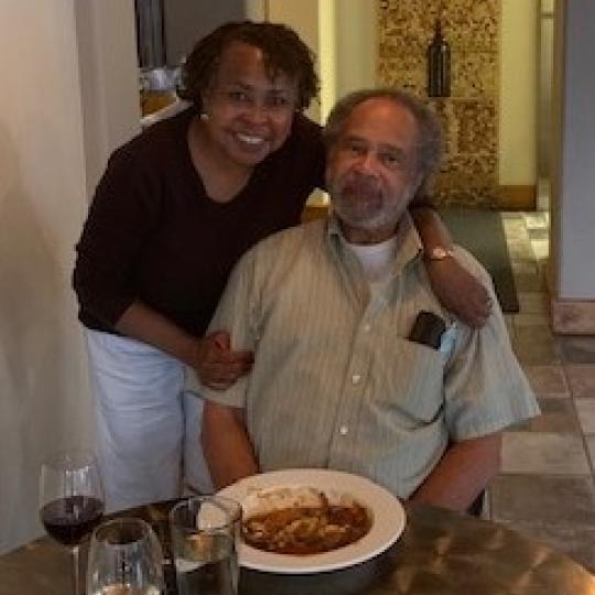 Photo of a man and a woman in a restaurant. He is sitting at a small table, upon which a bowl of pasta, as well as glasses of wine and water are placed. The woman is standing behind the seated man, with her arms around his shoulders. They are smiling.