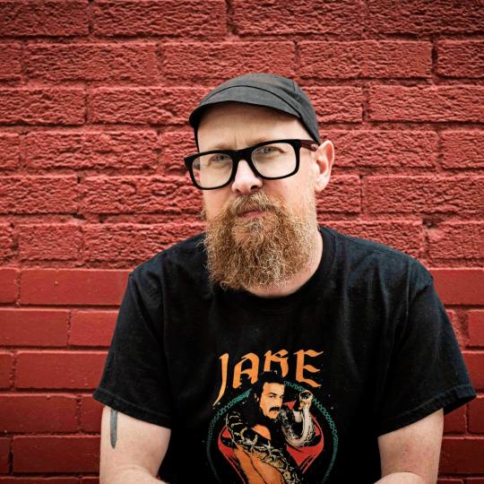 Photo from mid-chest up, of a man sitting in front of a brick wall that is painted reddish brown.  He is wearing a black cap with a narrow brim, and he has heavy black-framed eyewear and a curly brown beard of medium length. He is wearing a black t-shirt with the image of a moustached man, with the word "JAKE" at the top of the image. Part of a tattoo peeks out from underneath Chris' right short sleeve.