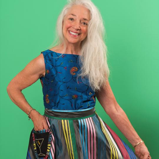 Photo of EllynAnne Geisel. She is standing in front of a bright green background. She has long silver blonde hair and she is smiling, wearing a deep blue sleeveless dress and  brightly colored striped apron. While her right hand rests on her right hip, her left hand is holding the left edge of the apron out so that the decoration is more visible.