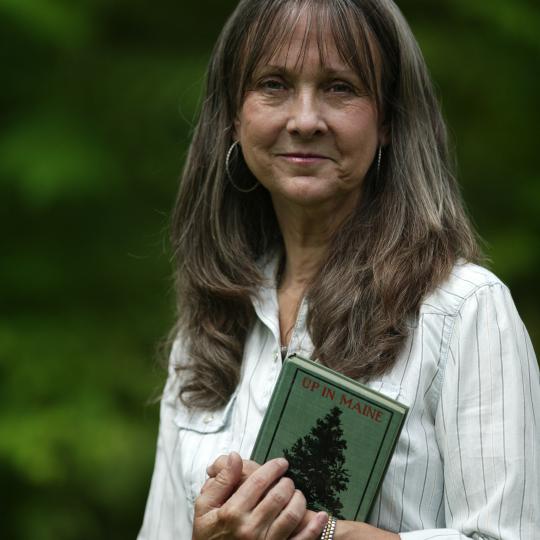 Photo of author Kayann Short. She is standing outdoors, green trees in the distance behind her. She has long, straight brown hair, and wears large silver hoop earrings and a white dress button-up blouse--the sleeves are rolled up to 3/4 length. Her hands are crossed in front of her chest as she holds onto a small hardcover book with a green cover, called "Up in Maine."