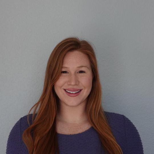 Photo of a young woman standing in front of a plain white wall for a portrait. She is wearing a scoop neckline turquoise colored shirt with long sleeves. She has long wavy red hair that falls below her shoulders, and lots of freckles. She is looking directly at the camera and smiling.