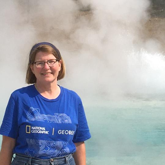 Photo of a woman standing in front of a geothermal pool. She is wearing a bright blue short sleeved t-shirt and has her shoulder length hair pulled back with a headband. She is smiling.