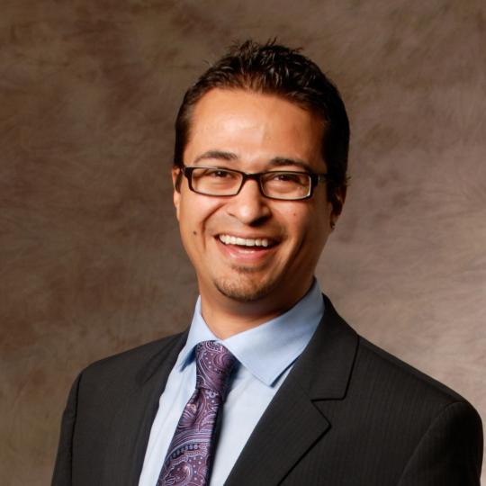 Photo of a man wearing a light blue dress shirt, purple and blue paisley necktie, and dark colored suit jacket. He is smiling. He has short cut dark hair and a small patch of facial hair on his chin. He is smiling, and he wears rectangular shaped eyewear.