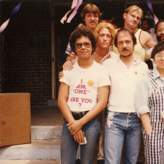 Seven individuals in 1970s clothing and fashion stand in front of a doorway, huddled close together and smiling. A man in the front row wears a shirt reading "I am 'One'. Are You?"