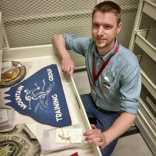Photo of a man, squatting down next to a collections drawer in a storage room with white shelves. His hands are resting on the drawer front edges, and he is looking at the cameral. The artifacts in the drawer are tagged and wrapped for storage. The largest artifact in the drawer is a blue sign shaped like a mountain with a white snow capped peak, which says, "Mountain Training Group." The man in the photo wears a blue button-collar shirt and blue jeans, with an ID badge on a lanyard around his neck.