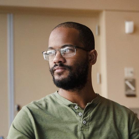Photo of Dexter Nelson II, an African American man wearing a green shirt and metal framed eyewear. He has close-cropped black hair, beard, and moustache. He is looking into the distance off to his right.