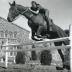A woman on a horse leaping a low barrier. A stable is visible behind them.