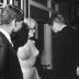 Photo of Robert Kennedy on the left of the image, standing next to Marilyn Monroe who is wearing the iconic nude-looking dress that is covered in rhinestones. President Kennedy stands next to her with his back to the camera and they are in a room with shelves filled with books. There is a partial image of a man captured on the right edge of the image. They are talking and smiling.
