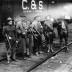 Four men standing on rail road tracks in front of a box car labelled C&S. The men are wearing hats and holding rifles.